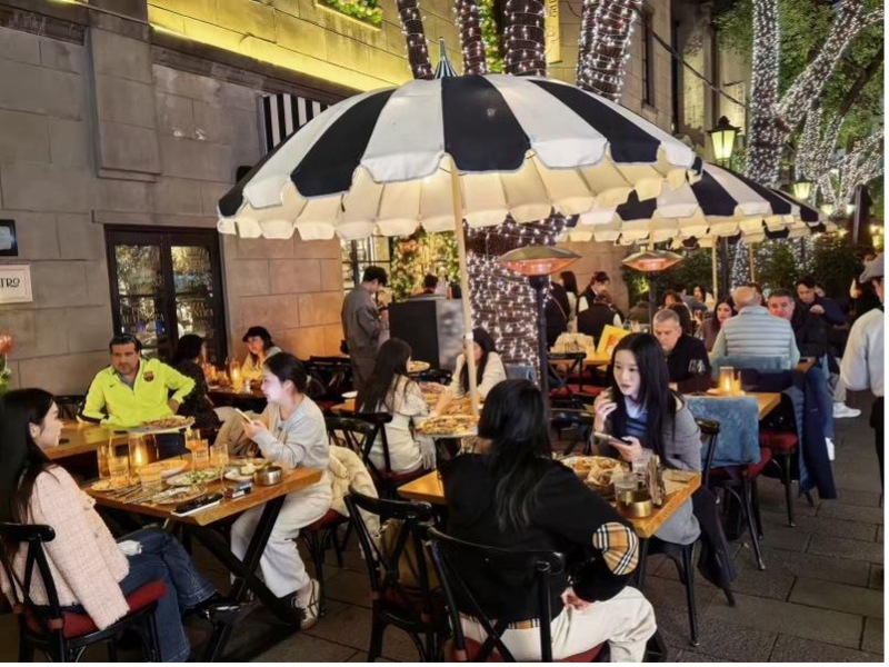 Background and ChallengeShanghai, as a global metropolis, faces the challenge of balancing commercial vibrancy with urban management. Outdoor dining (外摆位) has historical roots in Shanghai, but strict regulations on street vending and outdoor commercial activity, including bans on extending business operations beyond shop premises, had long constrained business vitality. In December 2022, Shanghai amended its Municipal Appearance and Environmental Sanitation Regulations. With respect to outdoor dining, the Regulations changed the relevant wording from “prohibited” to“prohibited without authorization,” which opened new possibilities for regulated outdoor dining. Since then, Shanghai has been continuously refining policies and service ecosystems, exploring efficient governance models to boost outdoor dining and the nighttime economy. In accordance with the municipal framework, district governments have updated their policies in this area to better manage and facilitate the orderly development of street vending and outdoor dining. Huangpu district, for example, issued version 2.0 of its outdoor dining policy in February 2026, expanding eligible locations for outdoor seating and permits extended operating hours for participating businesses, including bars and cafes. This policy development signals an acceleration in the development of Shanghai’s outdoor dining and nighttime economy.Policy TimelineThe revised Shanghai Municipal Appearance and Environmental Sanitation Regulations took effect on December 1, 2022, providing the legal foundation for subsequent reforms to allow greater flexibility for outdoor dining. Building on this, the Guidelines on Further Regulating Street Vending Activities (Trial), published on July 5, 2023, specifies that landmark nightlife areas, cultural districts, commercial circles, and scenic spots may permit time-based outdoor dining accounting for festivals and seasonal changes. Measures on Further Supporting the Development of Popular Nighttime Economy Landmarks were then jointly published on August 21, 2025 by the Shanghai Commission of Commerce and seven other Shanghai municipal authorities. The Measures support outdoor dining in areas of local landmarks during nighttime and aim to develop unique scenes of “outdoor dining economy.” In February 2026, the Huangpu District of Shanghai released two revised policy guidelines for street vending activities and outdoor dining. The two guidelines aim to create “outdoor dining–friendly neighborhoods” that attract both residents and tourists for leisure and consumption.Innovative ApproachDaxue Road of the city’s Yangpu District demonstrates a robust and innovative approach to outdoor dining management. The Road’s management authority led the establishment of an independent committee in 2019 that brings together the government, universities, enterprises, and local residents to jointly contribute to outdoor dining management in the neighbourhood. Since 2023, management standards have been developed and upgraded to version 2.0, adopting a “one road, one plan; one store, one standard” approach. These standards specify permitted zones and areas, unify facility materials, styles, and colors, and establish transparent approval processes with property and multi-department assessments. Accountability is enforced through formal self-governance guidelines: violations can result in loss of outdoor seating privileges, and serious or repeated infractions may lead to non-renewal of business leases. The 700-meter street now hosts nearly 80 specialty merchants, with outdoor seating increasing some businesses' revenue by approximately 30%. To address weather concerns, transparent wind shields were added in winter 2024, boosting sales by 24% from December to March, with February recording nearly 40% growth.The Measures issued in August 2025 facilitate the setup of outdoor seating at popular nighttime landmarks in Shanghai, add temporary outdoor seats for major festivals and simplify approval procedures for nighttime events. It also adds temporary nighttime parking spaces and pick-up points and promote time-based free parking areas. It encourages nighttime tourist bus routes that connect landmark buildings and nighttime economy sites, upgrades regional nighttime landscape lighting with 24-hour window displays lighting to create a unique commercial atmosphere.Additionally, the two latest policy documents released by Huangpu District in 2026 relax regulations on outdoor dining, including expanding eligible locations (such as permitting it in certain historical areas), extending operating hours for businesses with outdoor seating, and streamlining application procedures for individual businesses to establish outdoor seating areas.OutcomesAccording to the 2025 China Urban Nighttime Economy Index released by CBN and Rising Lab, Shanghai ranked first nationally with 4.03 million active nighttime outings in 2024. Since June 2025, nighttime consumption reached 88.09 billion yuan, up 3.3% year-on-year. In late July 2025, Shanghai designated its first batch of five “Nighttime Economy Popular Landmarks” and five “Nightlife Destinations.”Authors:Yacheng Sun is Professor of Marketing, School of Economics and Management, Tsinghua University.Maggie Wenjing Liu is Associate Professor of Marketing, School of Economics and Management, Tsinghua University.Lesley Luyang Song is Associate Professor of Marketing, School of Management,Huazhong University of Science and Technology.
